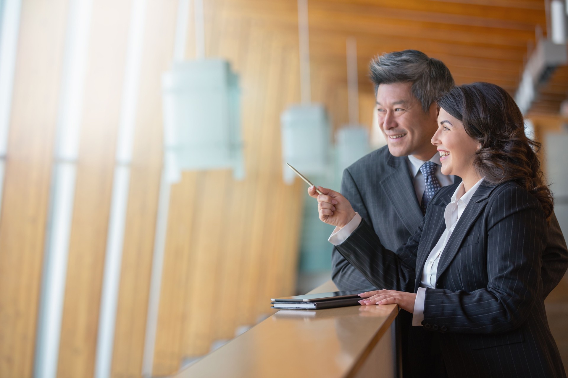 Mature Caucasian businesswoman and Chinese Asian businessman standing on office balcony taking notes and looking away